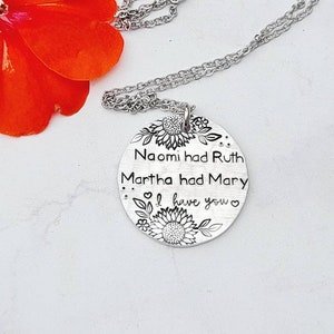 Może przedstawiać: Srebrny naszyjnik z wisiorkiem w kształcie okrągłej zawieszki. Na zawieszce wygrawerowano słowa "Naomi had Ruth, Martha had Mary, I have you" z motywami słonecznik&oacute;w i kwiat&oacute;w. Naszyjnik leży na białej powierzchni z czerwoną kwiatem.
