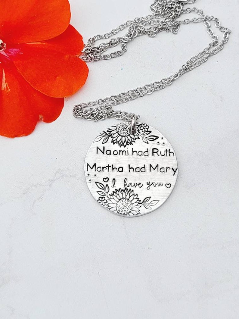 Może przedstawiać: Srebrny naszyjnik z wisiorkiem w kształcie okrągłej zawieszki. Na zawieszce wygrawerowano słowa "Naomi had Ruth, Martha had Mary, I have you" z motywami słonecznik&oacute;w i kwiat&oacute;w. Naszyjnik leży na białej powierzchni z czerwoną kwiatem.