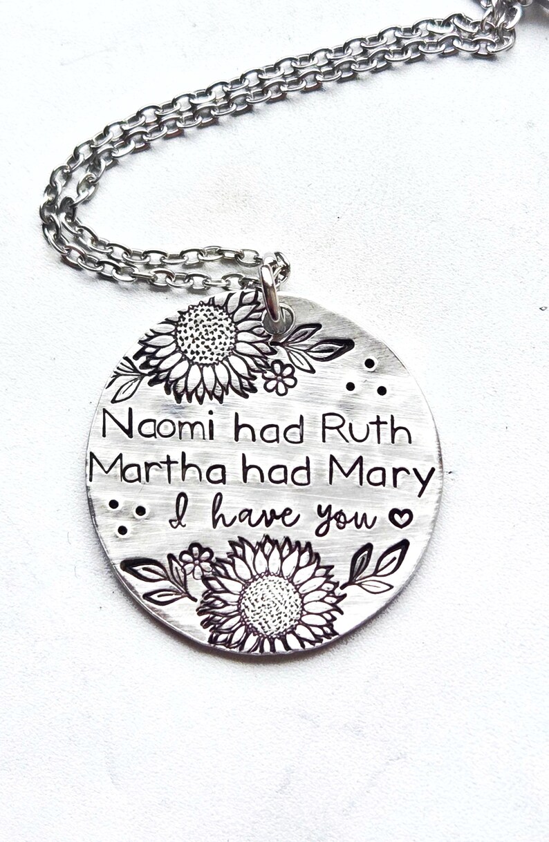 Może przedstawiać: Srebrny naszyjnik z okrągłą zawieszką. Zawieszka zawiera wygrawerowany tekst i ilustracje słonecznik&oacute;w. Tekst brzmi: "Naomi had Ruth, Martha had Mary, I have you".