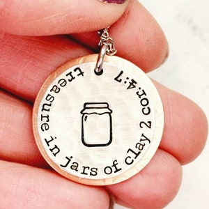 May include: A round pendant necklace with a silver chain. The pendant has a copper-colored outer ring and a silver-colored center. The center features a jar illustration and the text "treasure in jars of clay 2 cor. 4:7".