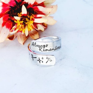 May include: A silver-colored wrap ring with the words "Always Remembered" engraved on the top. The ring also features dragonfly and star designs. A colorful flower with red, pink, and yellow petals is in the background.