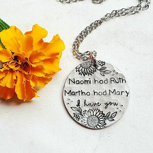 Może przedstawiać: Srebrny naszyjnik z wisiorkiem z okrągłym urokiem. Urok ma wygrawerowane słowa "Naomi had Ruth, Martha had Mary, I have you", z wzorami słonecznik&oacute;w i liści. Jasnopomarańczowy kwiat w tle.