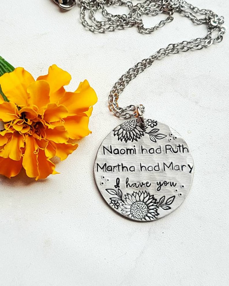 Może przedstawiać: Srebrny naszyjnik z wisiorkiem z okrągłym urokiem. Urok ma wygrawerowane słowa "Naomi had Ruth, Martha had Mary, I have you", z wzorami słonecznik&oacute;w i liści. Jasnopomarańczowy kwiat w tle.