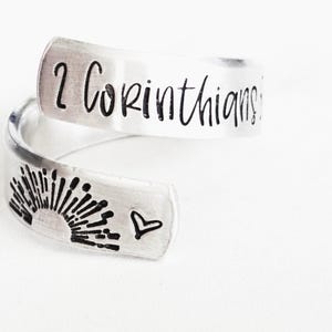 Puede incluir: Un anillo abierto de metal color plata con el texto grabado 2 Corinthians en la banda superior. La banda inferior muestra un gr&aacute;fico estampado de un sol naciente sobre una l&iacute;nea de horizonte, junto a un peque&ntilde;o s&iacute;mbolo de coraz&oacute;n.