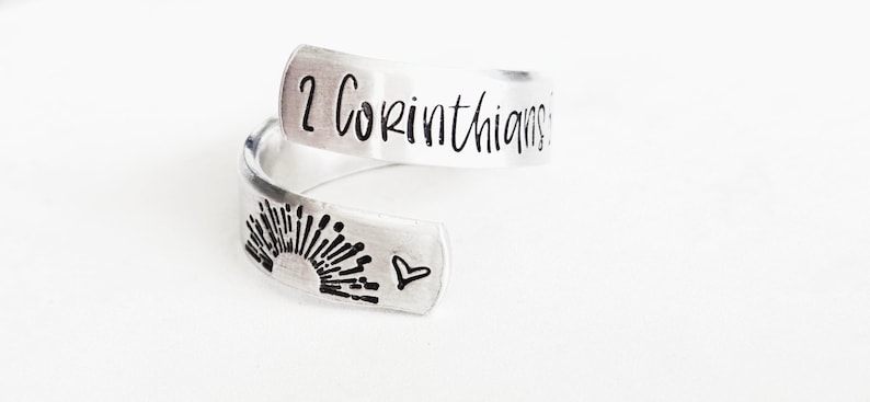 Puede incluir: Un anillo abierto de metal color plata con el texto grabado 2 Corinthians en la banda superior. La banda inferior muestra un gr&aacute;fico estampado de un sol naciente sobre una l&iacute;nea de horizonte, junto a un peque&ntilde;o s&iacute;mbolo de coraz&oacute;n.