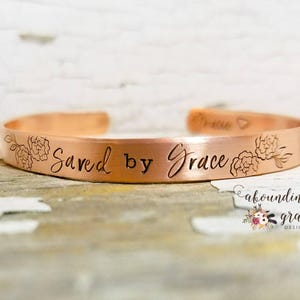 May include: A copper bracelet with the words "Saved by Grace" engraved on it. The bracelet is decorated with two small floral designs.