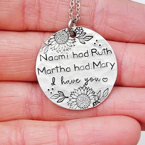 Może przedstawiać: Srebrny naszyjnik z wisiorkiem z okrągłą zawieszką. Zawieszka zawiera wygrawerowany tekst: &bdquo;Naomi miała Rut, Marta miała Marię, ja mam ciebie&rdquo;. Słoneczniki i liście zdobią g&oacute;rę i d&oacute;ł. Naszyjnik trzymany jest w dłoni.