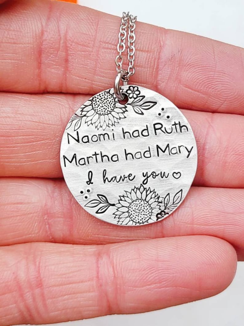 Może przedstawiać: Srebrny naszyjnik z wisiorkiem z okrągłą zawieszką. Zawieszka zawiera wygrawerowany tekst: &bdquo;Naomi miała Rut, Marta miała Marię, ja mam ciebie&rdquo;. Słoneczniki i liście zdobią g&oacute;rę i d&oacute;ł. Naszyjnik trzymany jest w dłoni.