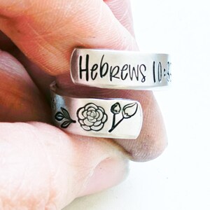 Puede incluir: Un anillo abierto de tono plateado con la inscripci&oacute;n Hebrews 10:35 estampada en la banda superior. La banda inferior muestra un dise&ntilde;o grabado en negro que consiste en una peque&ntilde;a rama con hojas, una rosa detallada y un capullo de flor cerrado.