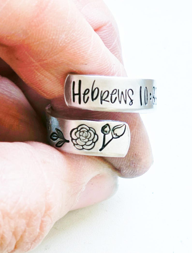 Puede incluir: Un anillo abierto de tono plateado con la inscripci&oacute;n Hebrews 10:35 estampada en la banda superior. La banda inferior muestra un dise&ntilde;o grabado en negro que consiste en una peque&ntilde;a rama con hojas, una rosa detallada y un capullo de flor cerrado.