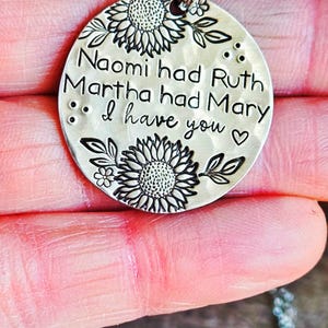 Może przedstawiać: Srebrny naszyjnik z okrągłą zawieszką. Zawieszka zawiera napis "Naomi had Ruth, Martha had Mary, I have you" otoczony wzorami słonecznik&oacute;w i liści. Naszyjnik jest trzymany w dłoni.