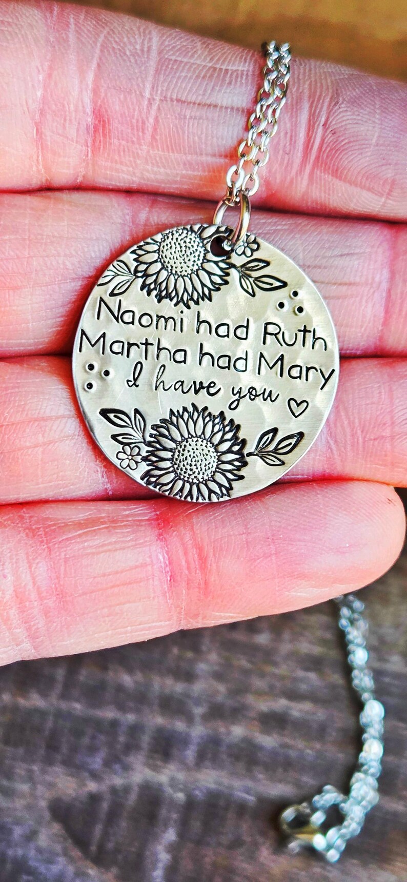 Może przedstawiać: Srebrny naszyjnik z okrągłą zawieszką. Zawieszka zawiera napis "Naomi had Ruth, Martha had Mary, I have you" otoczony wzorami słonecznik&oacute;w i liści. Naszyjnik jest trzymany w dłoni.