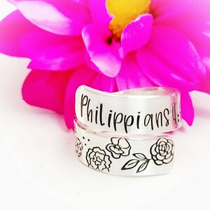 Puede incluir: Un anillo envolvente de tono plateado con el texto grabado Philippians 4:13 en la banda superior y una serie de ilustraciones florales de l&iacute;neas negras en la banda inferior, colocado frente a una flor rosa brillante con centro amarillo.
