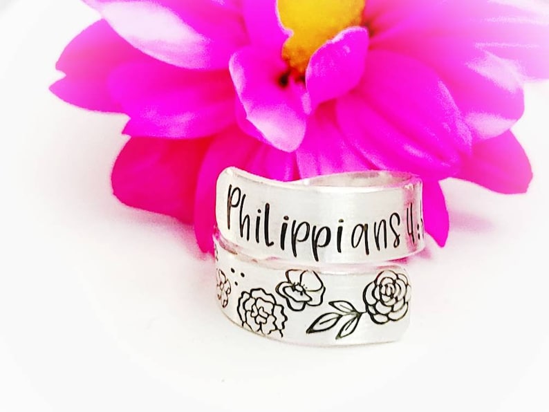 Puede incluir: Un anillo envolvente de tono plateado con el texto grabado Philippians 4:13 en la banda superior y una serie de ilustraciones florales de l&iacute;neas negras en la banda inferior, colocado frente a una flor rosa brillante con centro amarillo.