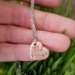 May include: A copper heart-shaped pendant necklace with a silver chain. The pendant has the words "so loved john 3:16" engraved on it.