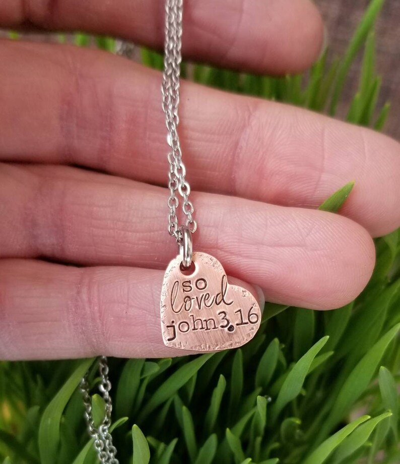 May include: A copper heart-shaped pendant necklace with a silver chain. The pendant has the words "so loved john 3:16" engraved on it.