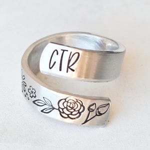 Puede incluir: Un anillo abierto de metal plateado con acabado cepillado. El anillo tiene grabadas las letras CTR en un extremo y una ilustraci&oacute;n detallada en l&iacute;neas negras de una rosa en flor con hojas y peque&ntilde;as flores a lo largo de la banda met&aacute;lica.