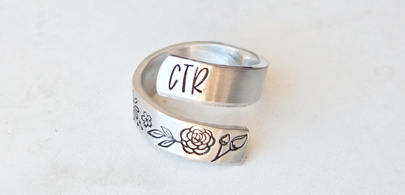 Puede incluir: Un anillo abierto de metal plateado con acabado cepillado. El anillo tiene grabadas las letras CTR en un extremo y una ilustraci&oacute;n detallada en l&iacute;neas negras de una rosa en flor con hojas y peque&ntilde;as flores a lo largo de la banda met&aacute;lica.