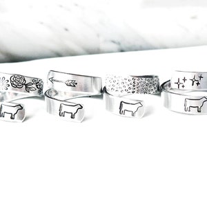 May include: A collection of silver-toned wrap rings. Each ring features a different design, including floral patterns, an arrow, a leopard print, stars, and a cow. The rings are displayed on a white surface.