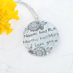 Może przedstawiać: Srebrny naszyjnik z okrągłą zawieszką. Zawieszka zawiera tekst "Naomi miała Rut, Marta miała Marię, ja mam ciebie" z motywami słonecznik&oacute;w i liści. Naszyjnik jest prezentowany na białej powierzchni z ż&oacute;łtym kwiatem.