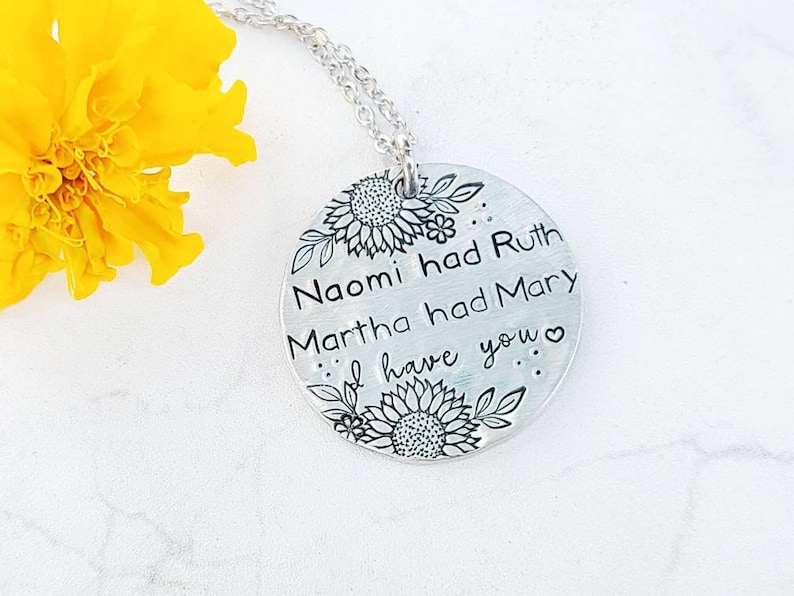 Może przedstawiać: Srebrny naszyjnik z okrągłą zawieszką. Zawieszka zawiera tekst "Naomi miała Rut, Marta miała Marię, ja mam ciebie" z motywami słonecznik&oacute;w i liści. Naszyjnik jest prezentowany na białej powierzchni z ż&oacute;łtym kwiatem.