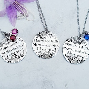 Może przedstawiać: Trzy srebrne naszyjniki z wisiorkami z motywami kwiatowymi i napisem "Naomi miała Rut, Marta miała Mary, Mam cię." Każdy naszyjnik ma inny kolorowy urok z kamieniem szlachetnym: czerwony, niebieski i r&oacute;żowy. Fioletowy kwiat jest w tle.