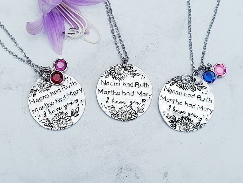 Może przedstawiać: Trzy srebrne naszyjniki z wisiorkami z motywami kwiatowymi i napisem "Naomi miała Rut, Marta miała Mary, Mam cię." Każdy naszyjnik ma inny kolorowy urok z kamieniem szlachetnym: czerwony, niebieski i r&oacute;żowy. Fioletowy kwiat jest w tle.