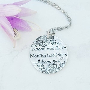 Może przedstawiać: Srebrny naszyjnik z wisiorkiem z okrągłą zawieszką z motywami kwiatowymi i napisem "Naomi had Ruth, Martha had Mary... I have you." Naszyjnik jest prezentowany na białej powierzchni z fioletowym kwiatem.