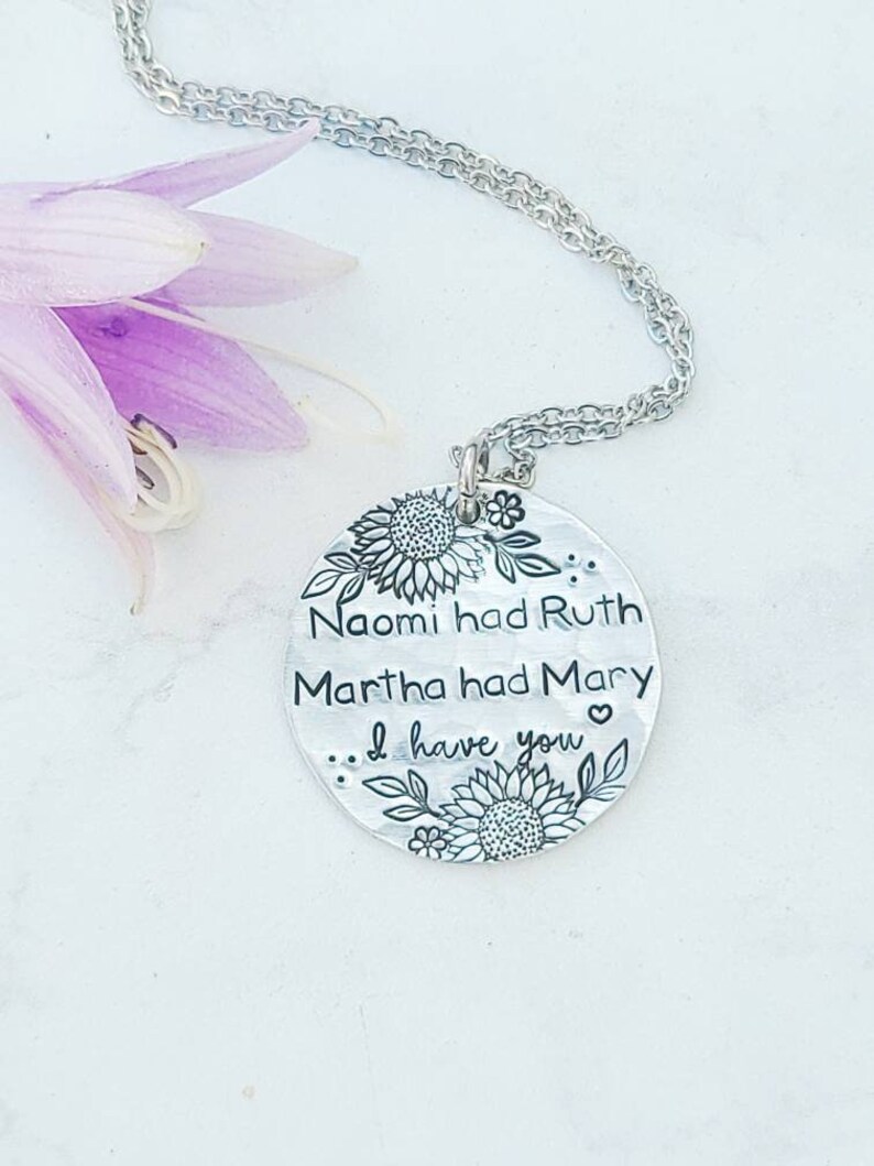 Może przedstawiać: Srebrny naszyjnik z wisiorkiem z okrągłą zawieszką z motywami kwiatowymi i napisem "Naomi had Ruth, Martha had Mary... I have you." Naszyjnik jest prezentowany na białej powierzchni z fioletowym kwiatem.