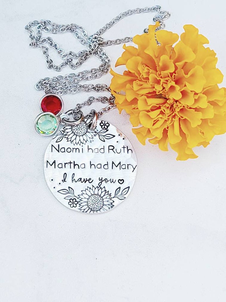 Może przedstawiać: Srebrny naszyjnik z okrągłą zawieszką z wygrawerowanym tekstem i kwiatowymi wzorami. Na zawieszce widnieje napis "Naomi had Ruth, Martha had Mary, I have you". Dwa urocze kamienie szlachetne w kolorze czerwonym i zielonym zwisają z łańcuszka. Ż&oacute;łty kwiat w tle.