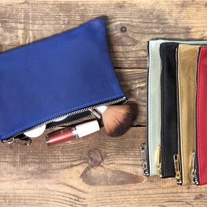 May include: Five zippered pouches in different colors, including blue, green, black, brown, and red. The pouches are made of leather and have a zipper closure. The blue pouch is open and contains makeup brushes and other items.