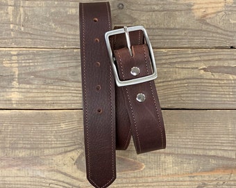 May include: A brown leather belt with a silver buckle. The belt is folded in half and the buckle is visible in the center of the image.