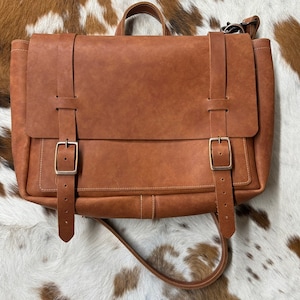 May include: A brown leather messenger bag with two adjustable straps and a buckle closure. The bag has a flap with a buckle closure and a top handle.