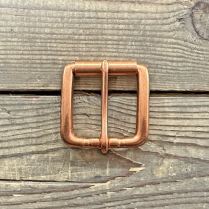 May include: A copper-colored belt buckle with a square shape and a smooth finish.