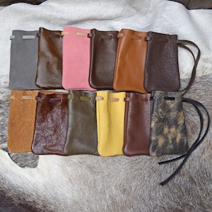 May include: Assortment of 10 leather pouches in various colors, including brown, gray, green, pink, and yellow. The pouches have a drawstring closure and are lined up on a white fur surface.