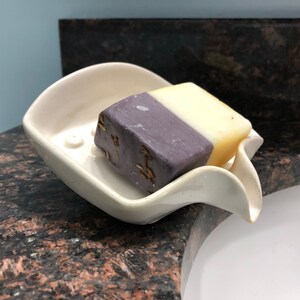 May include: A white ceramic soap dish with a curved design holds a bar of soap. The soap is half purple and half yellow.