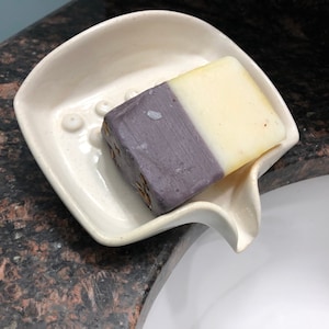 May include: A white ceramic soap dish with a curved spout and a raised design of small circles. A bar of soap with a purple and white colour scheme is resting in the dish.