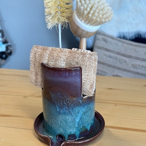May include: A handmade ceramic sponge holder with a dark purple and blue glaze. It holds a natural loofah sponge, a dish brush, and a bottle brush. The holder sits on a matching tray with a small spout.