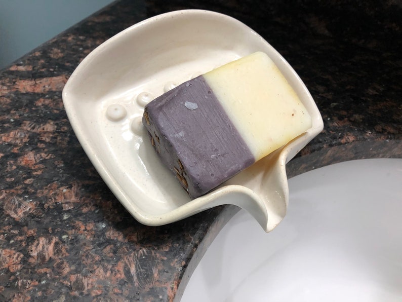 May include: A white ceramic soap dish with a curved spout and a raised design of small circles. A bar of soap with a purple and white colour scheme is resting in the dish.