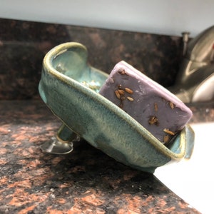 May include: A light blue ceramic soap dish with a suction cup base, holding a purple bar of soap with dried lavender flowers embedded in it.
