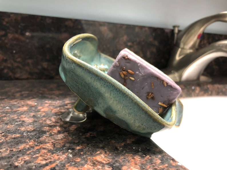 May include: A light blue ceramic soap dish with a suction cup base, holding a purple bar of soap with dried lavender flowers embedded in it.