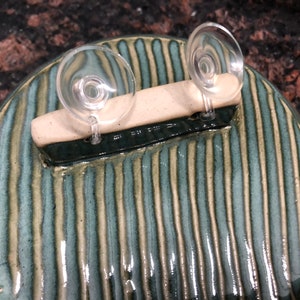 May include: A round ceramic dish with a green and white striped pattern. Two clear suction cups are attached to a white bar on top of the dish.