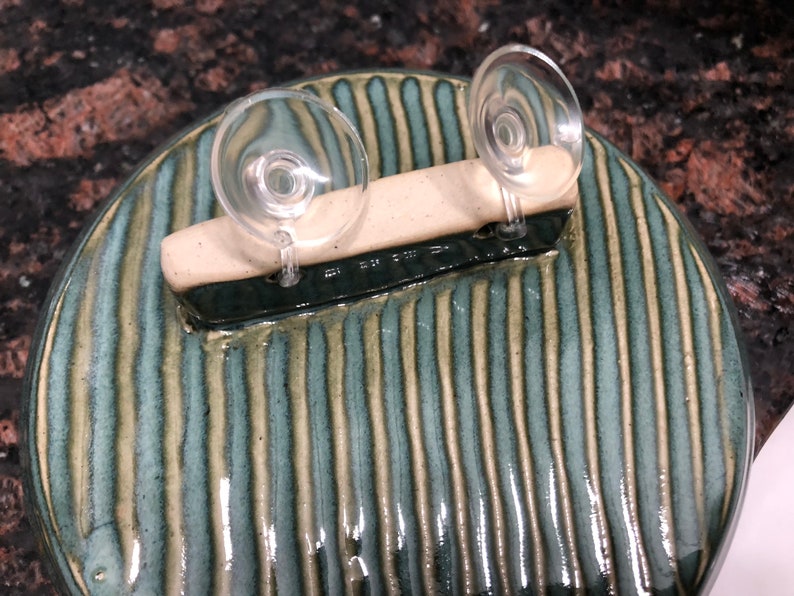 May include: A round ceramic dish with a green and white striped pattern. Two clear suction cups are attached to a white bar on top of the dish.