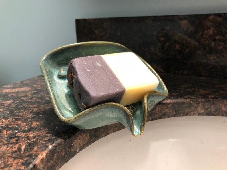 May include: A teal ceramic soap dish with a curved spout, holding a bar of soap with a purple and white design.