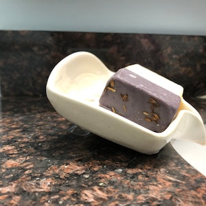 May include: A white ceramic soap dish with a suction cup on the bottom. A bar of purple soap with gold flecks is in the dish.