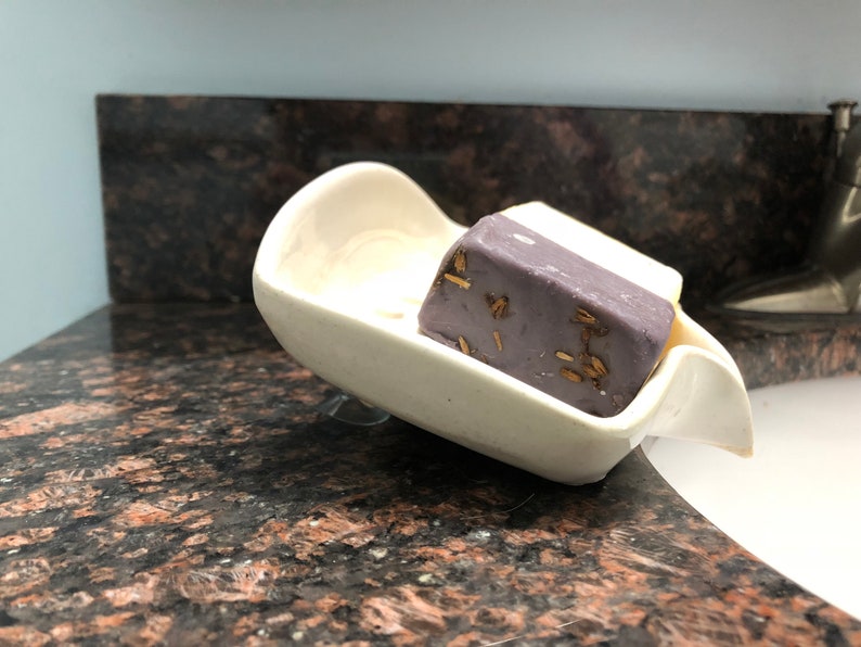 May include: A white ceramic soap dish with a suction cup on the bottom. A bar of purple soap with gold flecks is in the dish.