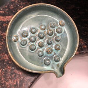 May include: A teal ceramic soap dish with a textured surface and a drain hole. The dish has a pattern of raised circles.