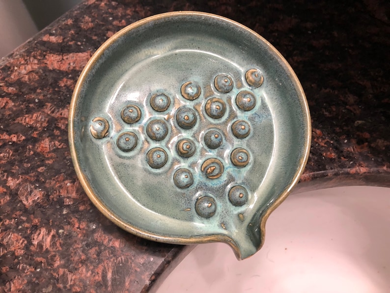 May include: A teal ceramic soap dish with a textured surface and a drain hole. The dish has a pattern of raised circles.