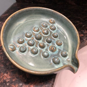 May include: A teal ceramic soap dish with a textured surface and a drain spout. The dish has a pattern of raised circles.