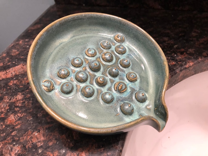 May include: A teal ceramic soap dish with a textured surface and a drain spout. The dish has a pattern of raised circles.